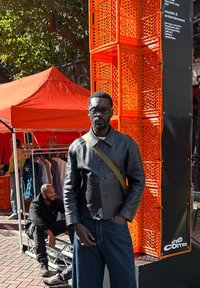 Sort læderjakke med knaplukning, grøn skulderrem, blå jeans, sat mod en orange kassebaggrund og et rødt baldakin.