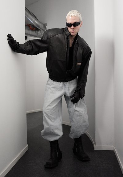 Persona con cabello corto y rubio que lleva gafas de sol negras, chaqueta de cuero negra, guantes negros, pantalones anchos gris claro y botas negras en un pasillo estrecho.