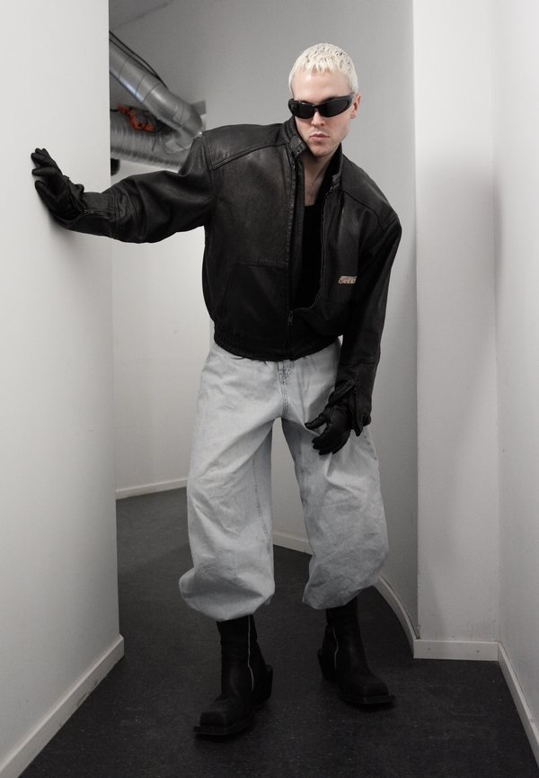 Person with short blond hair wearing black sunglasses, a black leather jacket, black gloves, loose light grey trousers, and black boots in a narrow hallway.
