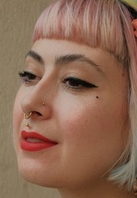 Nærbillede af et ansigt med pastelfarvet lyserødt hår, kraftig sort eyeliner, rød læbestift og flere guldpiercinger i næsen og på læben. Blød tekstur.