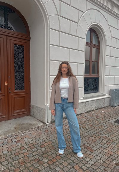 Chaqueta cropped de canalé marrón claro, camiseta blanca, jeans de cintura alta azul claro y zapatillas blancas, contra un muro de piedra texturizado.