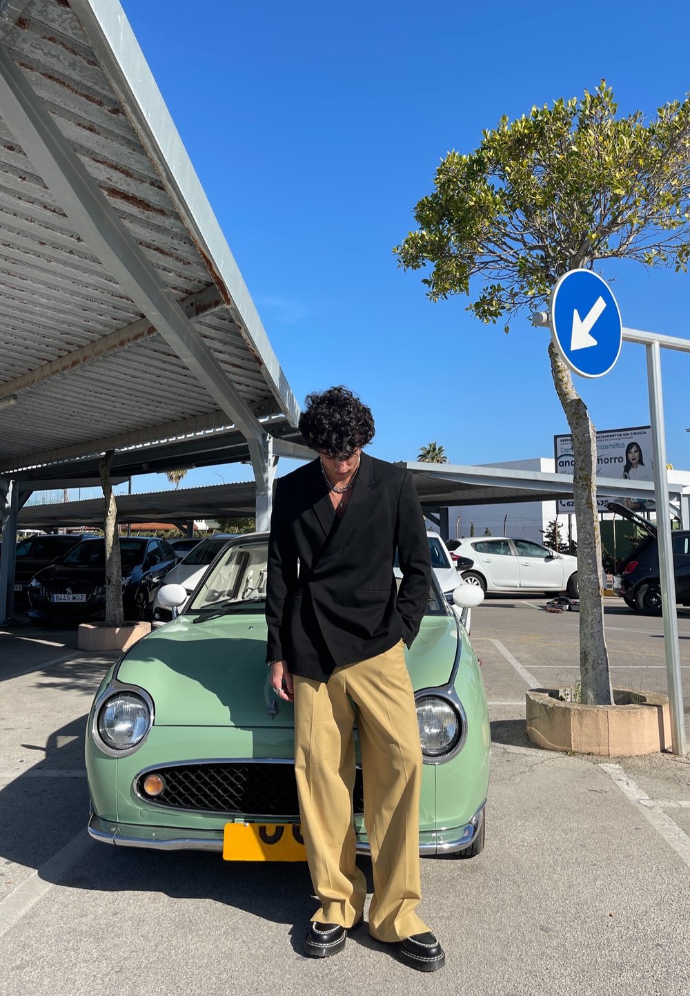 Blazer preto, calças largas bege, de pé ao lado de um carro vintage verde menta com faróis redondos e grelha de malha sob um céu azul.