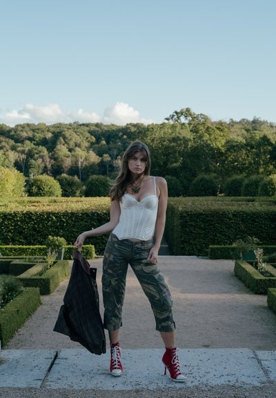 Top corsé blanco, pantalones cargo de camuflaje, botas rojas de tacón alto; sosteniendo una chaqueta oscura de cuadros en un entorno de jardín con vegetación.