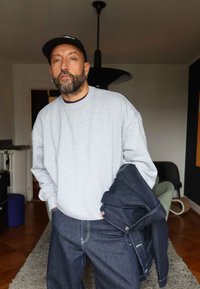 Grijze oversized sweatshirt met een ronde hals, gecombineerd met donkere denim broeken. Houdt een marineblauwe jas vast, voor een neutrale interieurachtergrond.