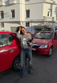 Czerwony samochód zaparkowany na ulicy, przy którym opiera się kobieta w swetrze w paski i dzwonach, trzymająca czarny przedmiot, światło słoneczne tworzy cienie.