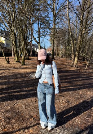 Joven con chaqueta azul claro, gorra rosa, pantalones vaqueros anchos y un bolso marrón camina por un sendero de parque cubierto de hojas a la luz del sol, con árboles sin hojas.