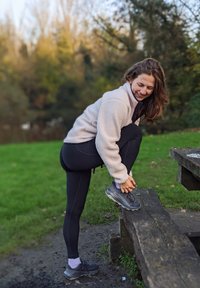 Pessoa veste uma jaqueta de fleece bege claro e leggings pretas, amarrando um tênis cinza enquanto se equilibra em um banco de madeira em um parque verde.
