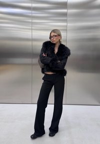 Sort faux fur jakke med en cropped design, parret med højtaljede sorte bukser og sorte sko, sat mod en metallisk baggrund.