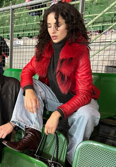 Chaqueta de cuero roja con un cuello de pelaje rojo, suéter negro de cuello alto, jeans azul claro, botas de cuero marrón y un bolso negro.