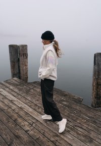 Veste en polaire crème avec motif zigzag pastel sur la manche, associée à un pantalon foncé et des baskets blanches, marchant sur une jetée en bois près de l'eau.