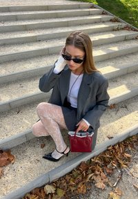 Grå oversized blazer, hvid skjorte med synlige manchetter, mønstrede hvide strømpebukser, sorte kitten heels og en lille rød håndtaske.