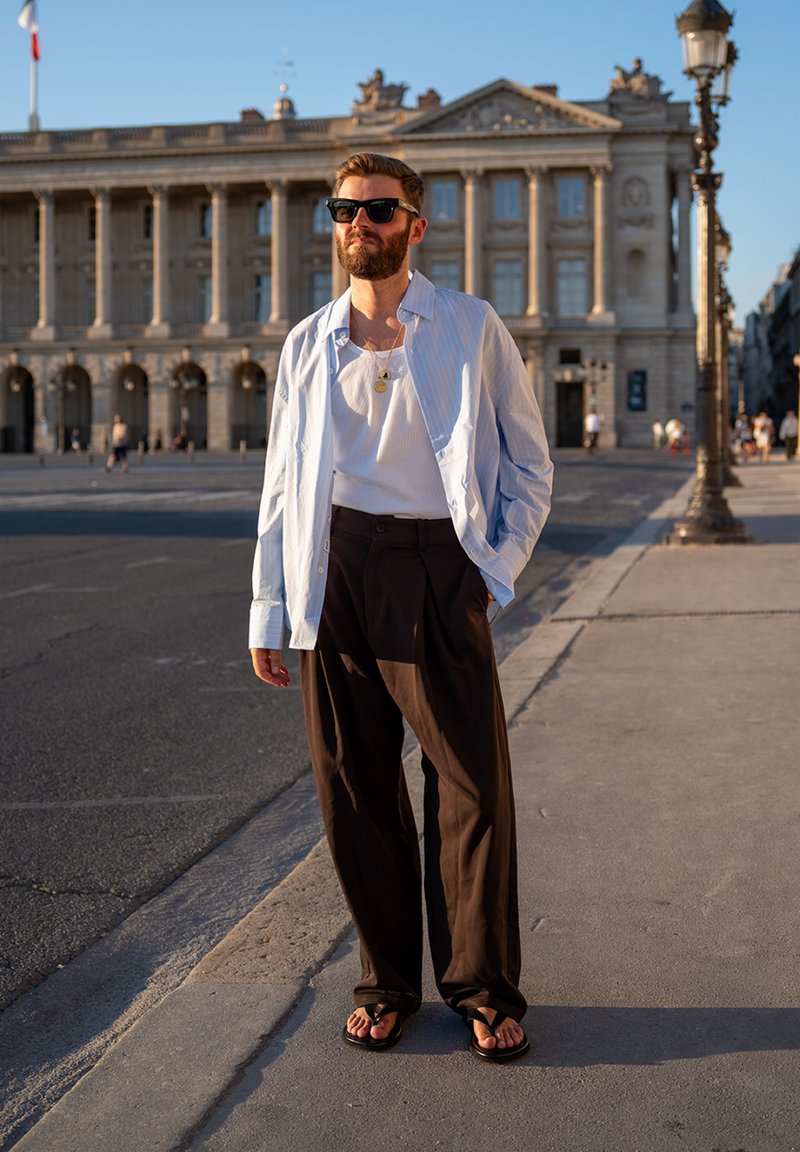 Homme portant des lunettes de soleil, chemise bleu clair, débardeur blanc, pantalon foncé et sandales, debout sur le trottoir de la ville près d’un bâtiment historique en plein soleil.