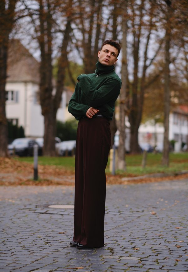 Pull en laine vert à col roulé avec une texture côtelée, associé à un pantalon bourgogne à jambes larges. Le modèle se tient sur un chemin en pavés dans un cadre extérieur.