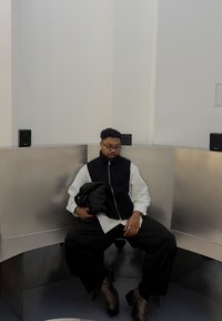 Fekete-fehér mellény hosszú ujjú ing felett, laza fekete nadrág és mintás barna csizma. Egy fém sarokban ül, hangszórók mellett.