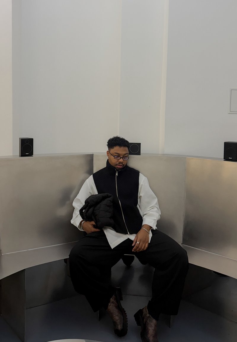 Zwart-wit vest over een shirt met lange mouwen, comfortabele zwarte broek en bruin patroonlaarzen. Zittend in een metalen hoek met luidsprekers.