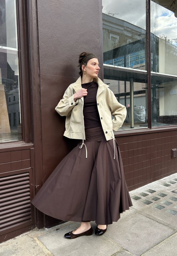 Femme appuyée contre un mur marron, portant une veste beige, un haut noir, une longue jupe brune et des ballerines noires, sur un trottoir en ville.
