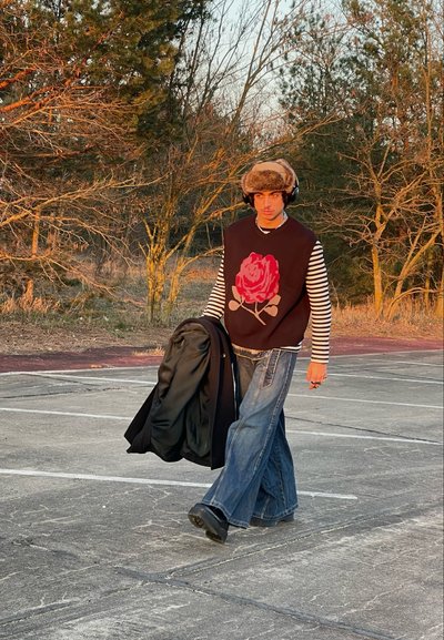 Chaleco negro con un gran gráfico de rosa, camisa de rayas de mangas largas debajo, vaqueros de pierna ancha y un sombrero forrado de piel. Lleva una chaqueta.