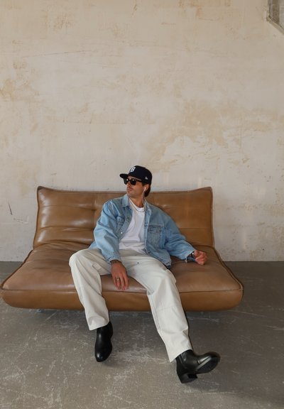 Hombre con gafas de sol, chaqueta vaquera, camisa blanca, pantalones blancos, botas negras y gorra sentado en un sofá de cuero marrón contra una pared texturizada.