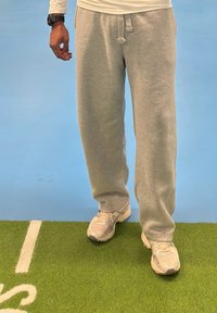 Personne portant un pantalon de survêtement gris et des baskets blanches debout sur un gazon synthétique vert à côté d'un sol bleu avec des marquages blancs.