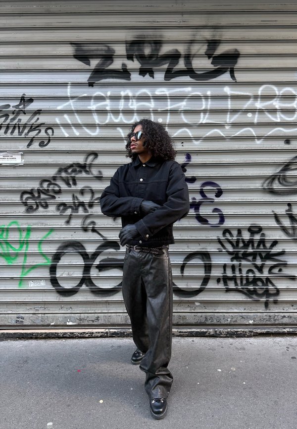 Person mit lockigem Haar, die schwarze Sonnenbrille, schwarze Jacke, Handschuhe und weite Lederhosen trägt, steht vor einem mit Graffiti bedeckten Metallrollladen.