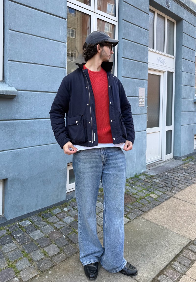 Homme portant une casquette noire, des lunettes de soleil, une veste marine, un pull rouge et un jean large bleu, debout dans une rue pavée devant un bâtiment bleu.