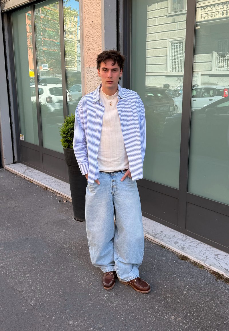 Νέος άνδρας που φοράει φαρδιά ανοιχτό μπλε τζιν, καφέ παπούτσια, λευκό πουκάμισο και μπλε ριγέ πουκάμισο στέκεται σε πεζοδρόμιο σε αστικό περιβάλλον κοντά σε κτίριο.