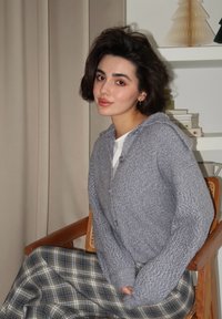 Cardigã cinza tricotado com capuz, apresentando um acabamento texturizado e fecho de botão. Combinado com uma saia xadrez em tons suaves.