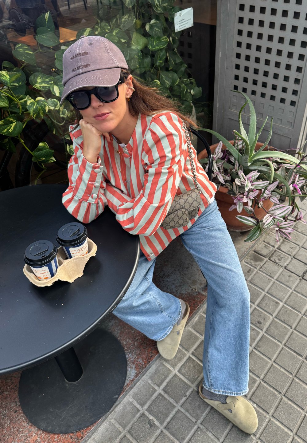Stribet rød og hvid skjorte, lyseblå brede jeans, beige klipklapper. Accessories inkluderer en grå baseball cap og en mønstret taske. To kaffekopper på bordet.