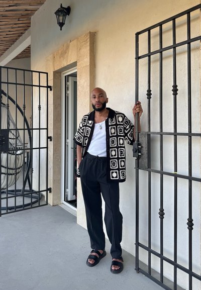 Camisa de overshirt en blanco y negro, camiseta blanca, pantalones negros y sandalias negras. De pie junto a una puerta de hierro forjado en un porche.