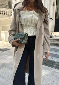 Μπεζ παλτό trench πάνω από ένα λευκό φουσκωτό crop top, συνδυασμένο με ψηλόμεσα σκούρα τζιν και μία υφασμάτινη τσάντα σε πράσινη απόχρωση.