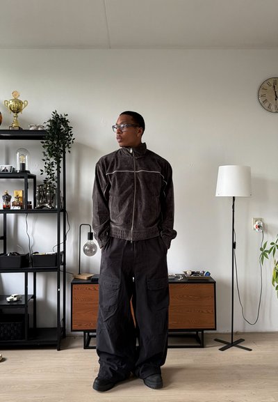 Hombre con chaqueta oscura con cremallera y pantalones negros anchos está de pie en una sala de estar con estanterías, una lámpara de pie y un reloj en la pared.