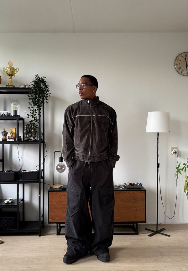 Man met een donker jasje met rits en wijde zwarte broek staat in een woonkamer met planken, een vloerlamp en een klok aan de muur.