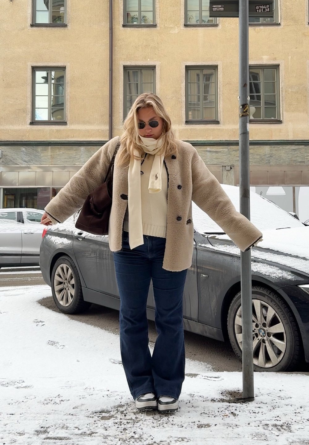 Casaco de peles bege com botões pretos, suéter creme, cachecol leve, jeans azuis à boca de sino e sapatilhas, de pé na neve ao lado de um carro cinza.