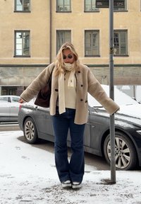 Casaco de peles bege com botões pretos, suéter creme, cachecol leve, jeans azuis à boca de sino e sapatilhas, de pé na neve ao lado de um carro cinza.