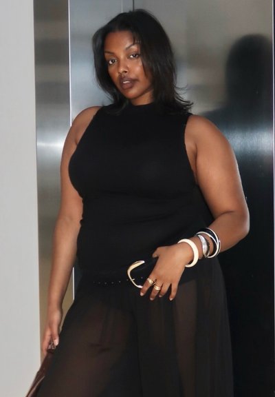 Mujer con top negro sin mangas y pantalones transparentes negros posa con pulseras y anillos frente a un fondo metálico.