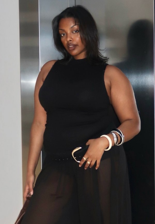 Femme en haut noir sans manches et pantalon noir transparent posant avec des bracelets et des bagues devant un fond métallique.