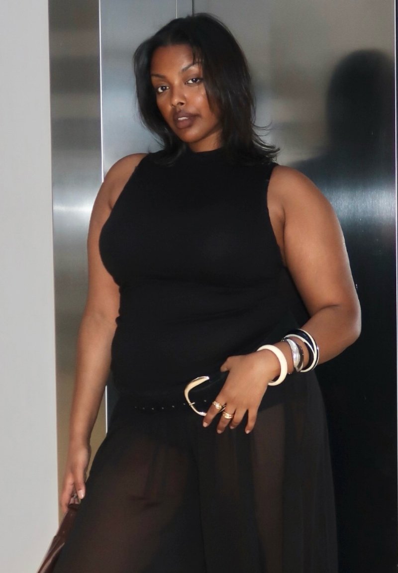 Kvinde i sort ærmeløs top og gennemsigtige sorte bukser poserer med armbånd og ringe mod metallisk baggrund.