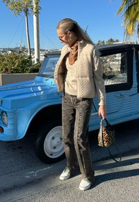 Femme aux cheveux blonds portant des lunettes de soleil, une veste en tricot beige, une écharpe marron, un jean foncé et des baskets blanches, debout à côté d'une voiture vintage bleue, tenant un sac à motif léopard.