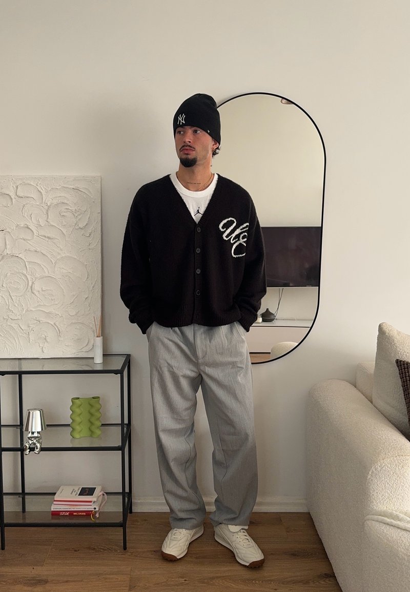 Μαύρο πλεκτό cardigan με λευκό λογότυπο, λευκό πουκάμισο, γκρίζα πλισέ παντελόνια και λευκά αθλητικά παπούτσια. Το φόντο περιλαμβάνει έναν καθρέφτη και διακοσμητικά αντικείμενα.