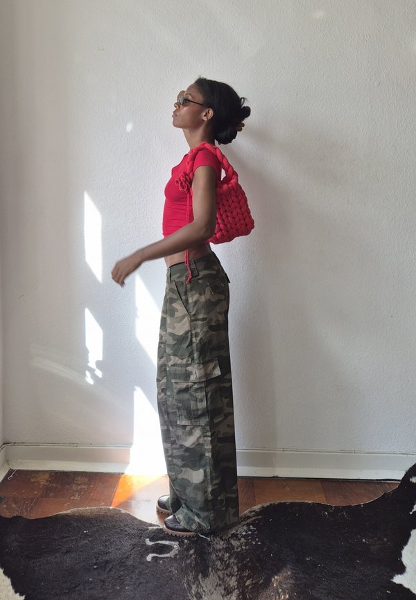 Woman wearing red top, camouflage pants, and glasses, carrying a chunky red knit bag on her shoulder, standing against a white wall with sunlight.