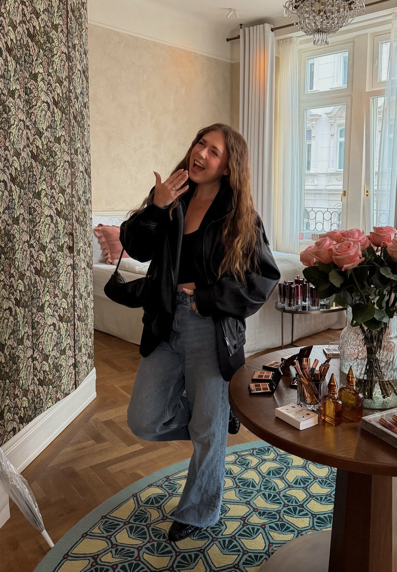 Jeune femme en veste noire et jean pose en souriant à l'intérieur près d'une table avec du maquillage et des roses roses près d'une fenêtre avec des rideaux blancs.