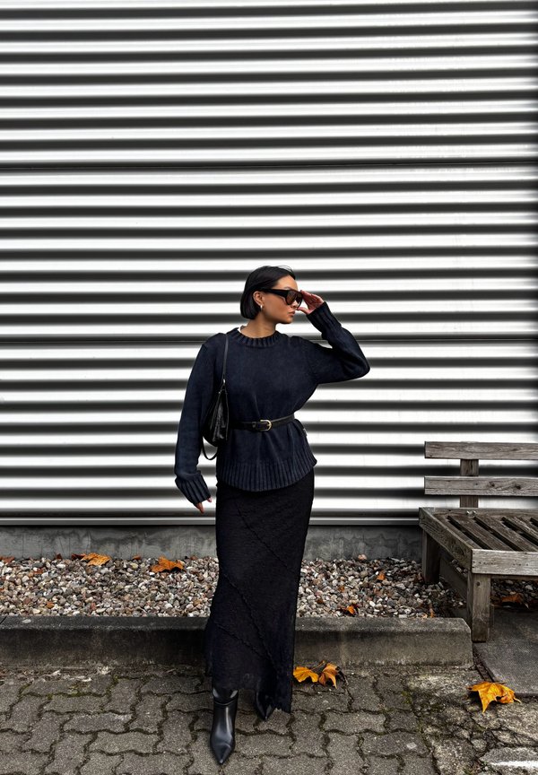 Schwarzer gestrickter Pullover mit geripptem Saum, kombiniert mit einem langen schwarzen Rock mit strukturiertem Muster. Accessoires umfassen einen schwarzen Gürtel und eine Tasche.