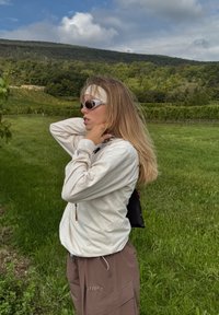 Osoba u svijetloj dugoj majici i smeškim cargo hlačama, s naočalama za sunce i trakom za kosu, stoji na travnatom polju.