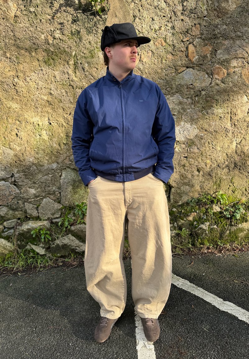 Hombre joven con chaqueta azul marino, pantalones anchos beige, zapatos marrones y gorra negra, de pie con las manos en los bolsillos frente a una pared de piedra.