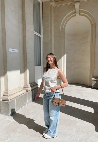 Conjunto informal que incluye una camiseta sin mangas de lino beige claro, jeans de pierna amplia azul claro y un pequeño bolso cruzado marrón. Entorno de piedra iluminado por el sol.