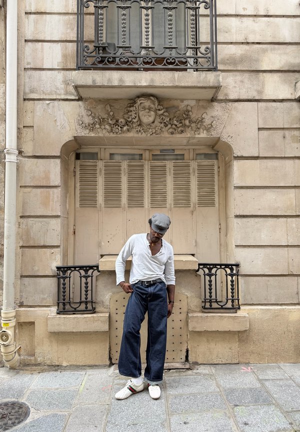 Hombre con camisa blanca, pantalones anchos azules y gorra gris de pie sobre un pavimento de piedra frente a un edificio beige con persianas cerradas y escultura decorativa de cara.
