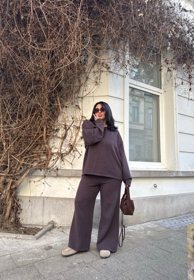 Mujer con suéter y pantalones morados de gran tamaño, gafas de sol y un pequeño bolso, de pie en la acera frente a un edificio con enredaderas secas.