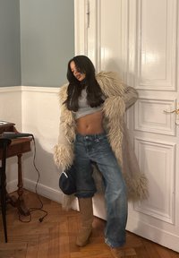 Long manteau en fausse fourrure beige avec de longs poignets duveteux, crop top gris, jean bleu ample et bottes montantes marron clair. En arrière-plan, une table et un mur.