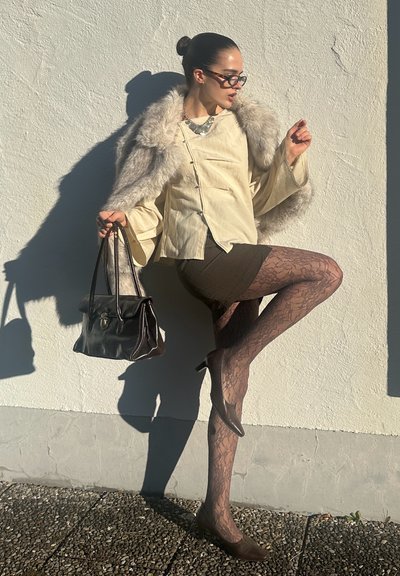 Blusa beige con detalles de piel, falda marrón, medias estampadas y tacones marrones; sosteniendo un bolso oscuro; sobre un fondo de pared blanca.