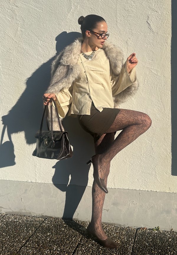 Beige blouse met bontaccenten, bruine rok, patroon panty's en bruine hakken; houdt een donkerkleurige handtas vast; tegen een witte muur gezet.